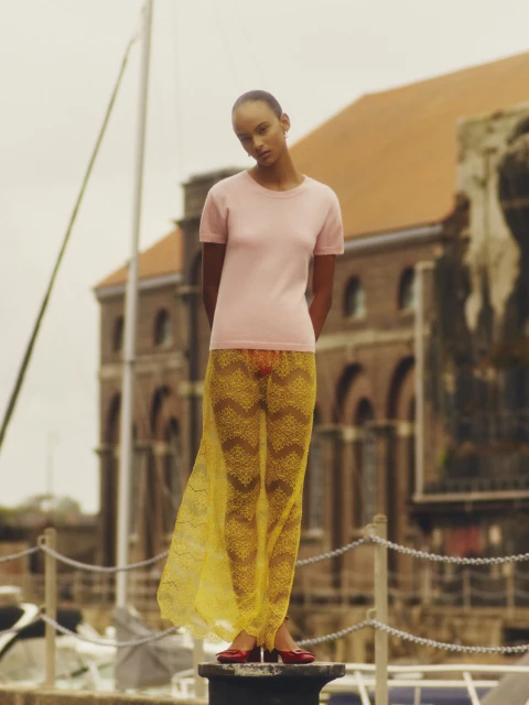 Fashion shot of a slender, light-skinned model in a pink top and yellow lace skirt posing on bollard with building in background.