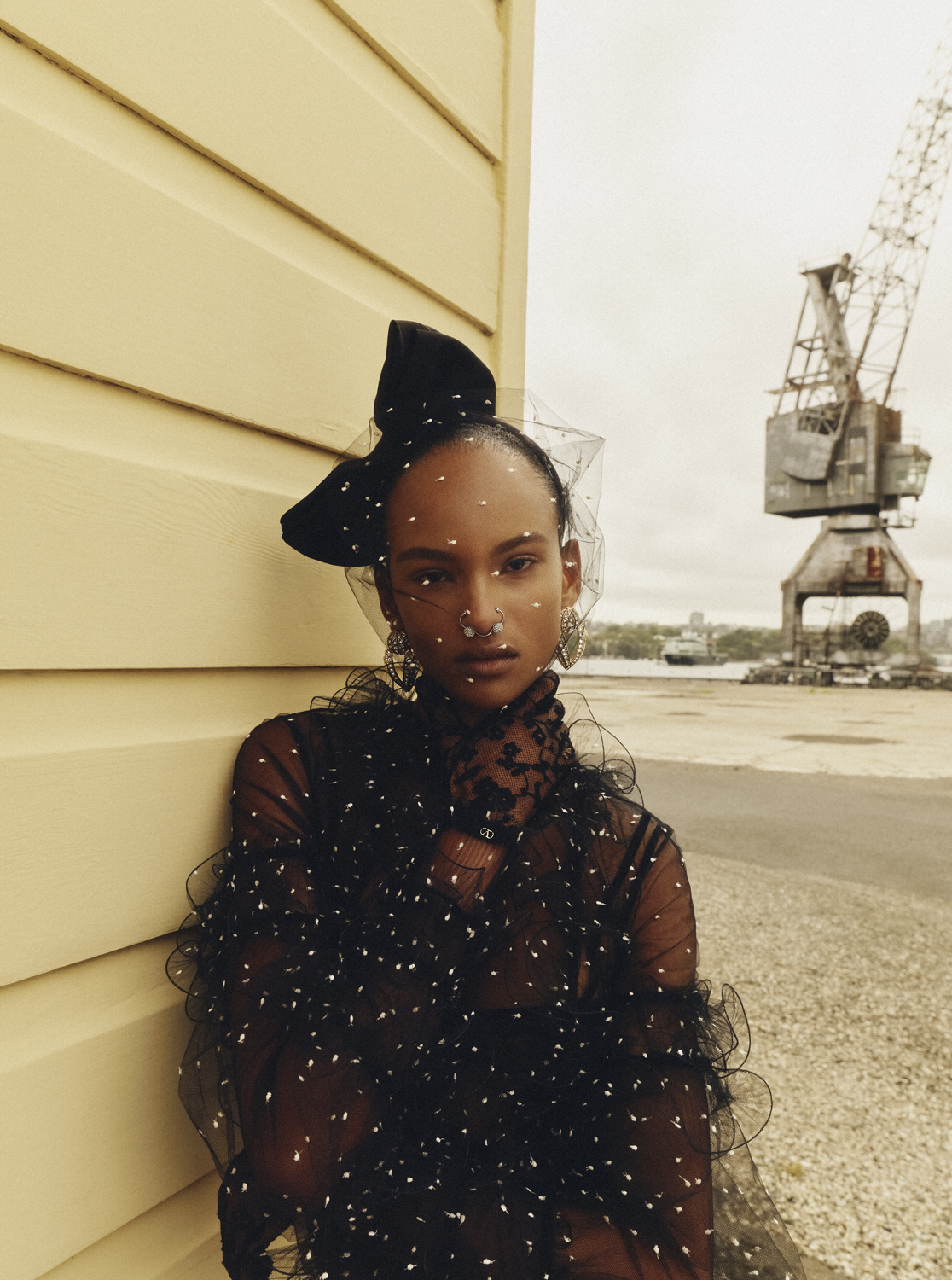 Portrait of a woman with a bow and veil, wearing a sheer top with embellishments, posing near a yellow wall and a crane.
