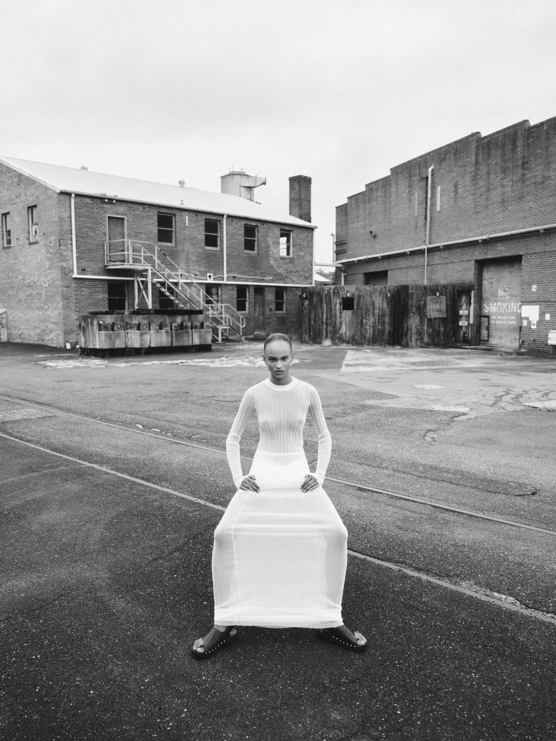 Monochrome fashion shot: a woman in a white dress poses outdoors in an industrial area, showcasing minimalist style against the backdrop of old brick buildings.