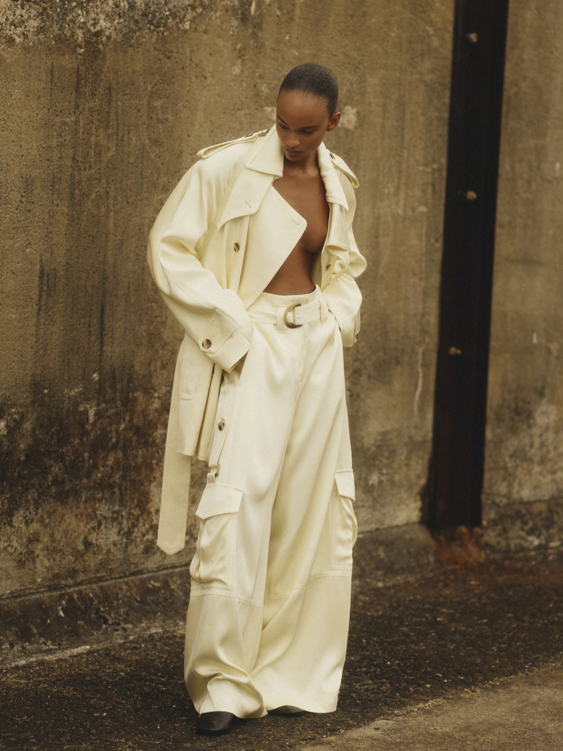 Woman poses in stylish, oversized cream-colored trench coat and wide-legged cargo pants against a textured, neutral-tone wall backdrop.