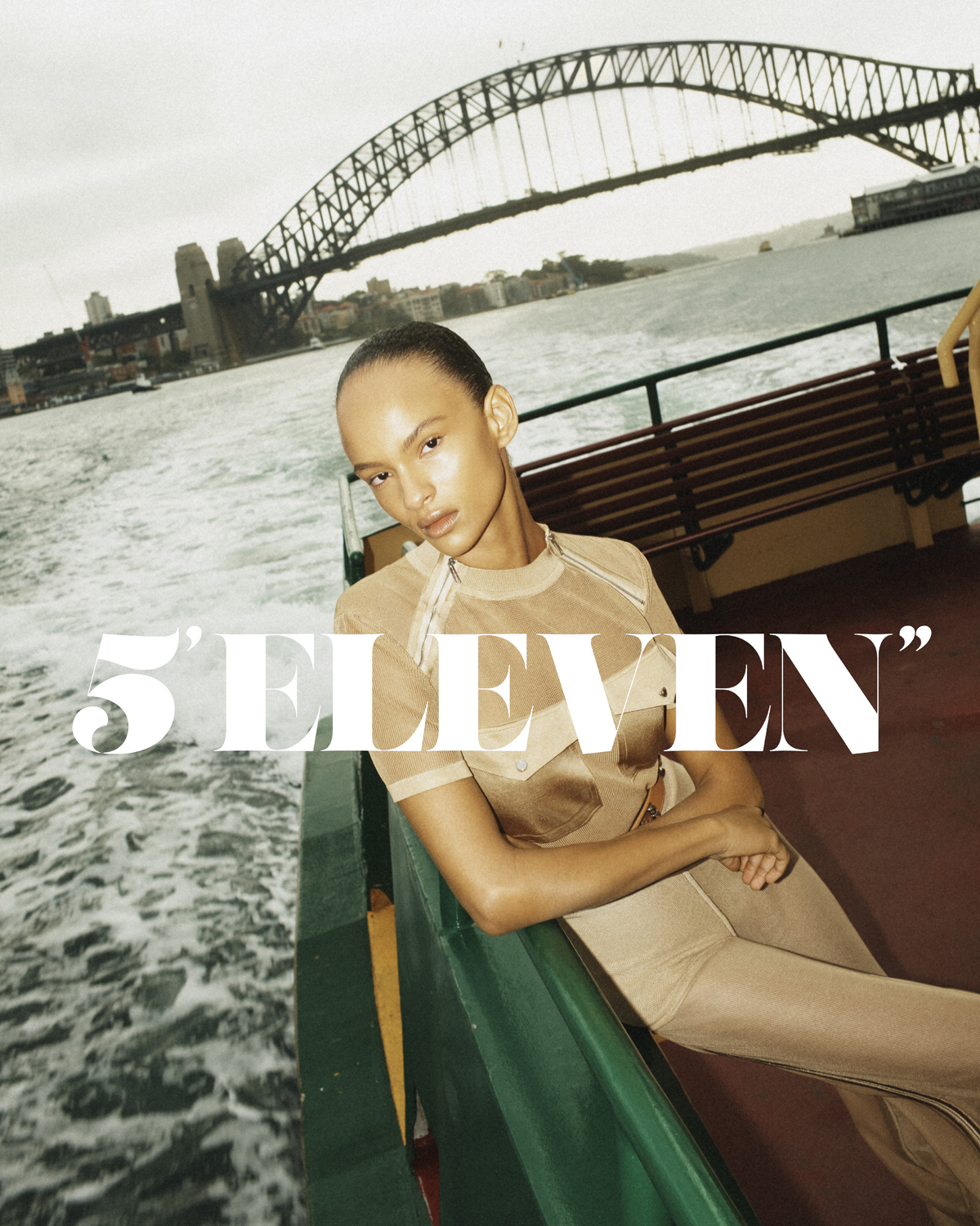 Fashion shot of a woman in beige clothing on a boat with a bridge in the background, the water reflects the overcast sky.