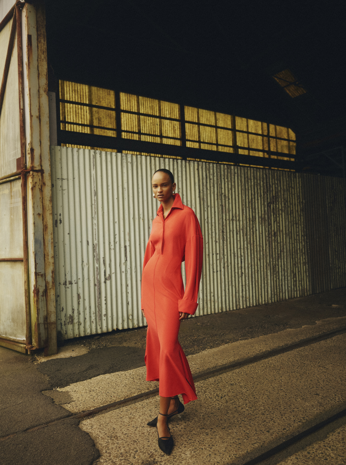 Adult woman in a red shirt-dress and black shoes poses on a street with a warehouse backdrop, captured in natural light.