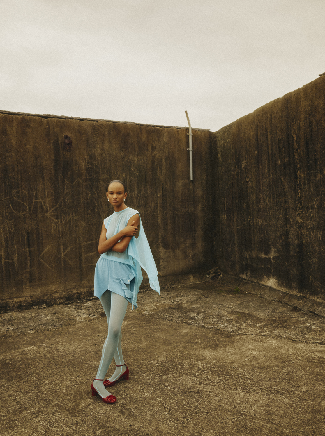 A single young adult woman with short hair is posed in a light blue dress and tights against a weathered, textured urban background.