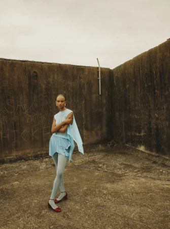 A single young adult woman with short hair is posed in a light blue dress and tights against a weathered, textured urban background.