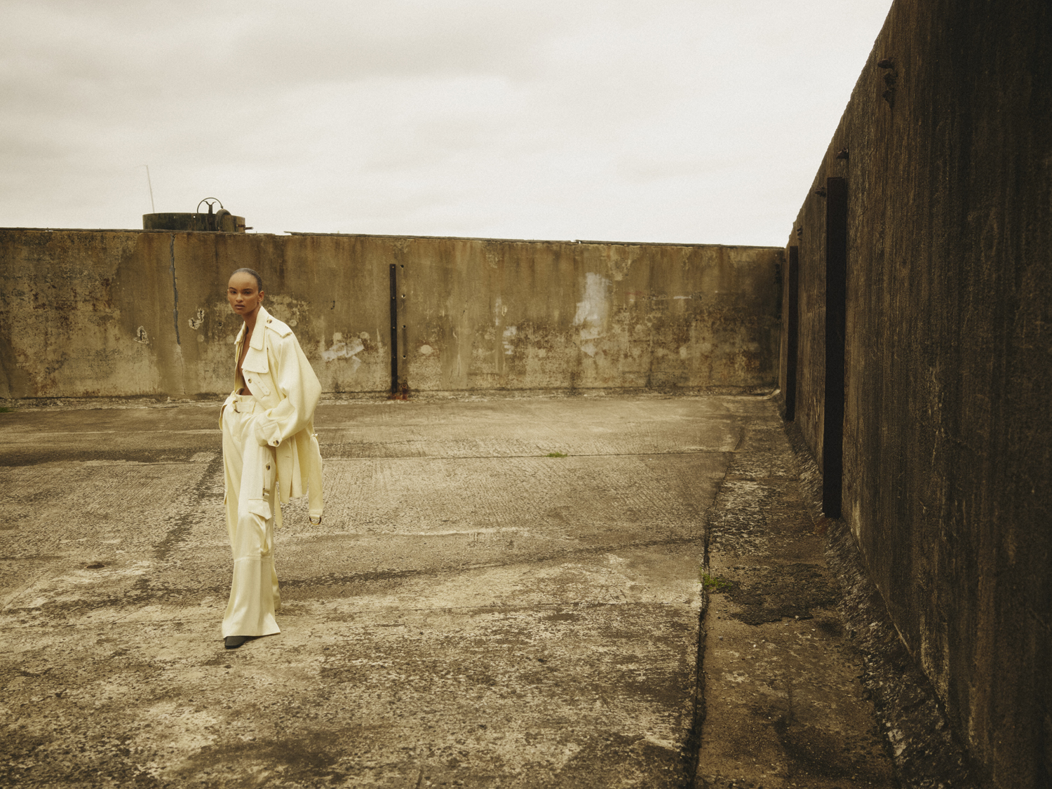 Fashionable woman in a trench coat and wide-leg pants poses outdoors on a weathered concrete surface against a cloudy sky.
