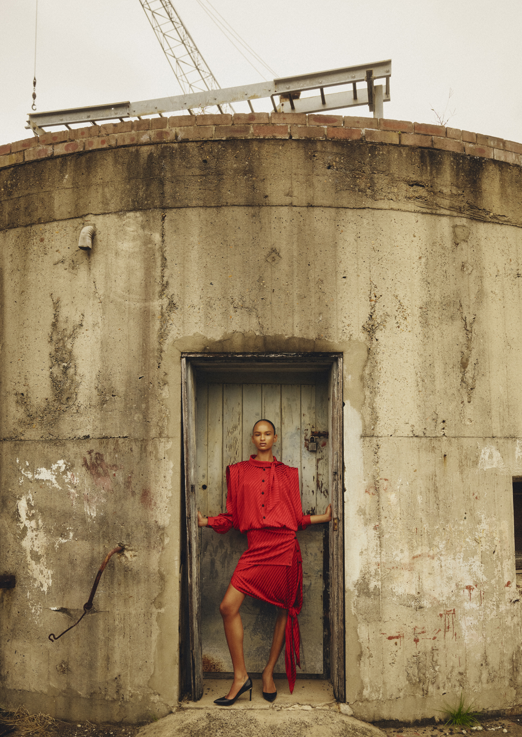 Fashionable woman in a red dress poses confidently in a weathered doorway with urban construction in the background on an overcast day.