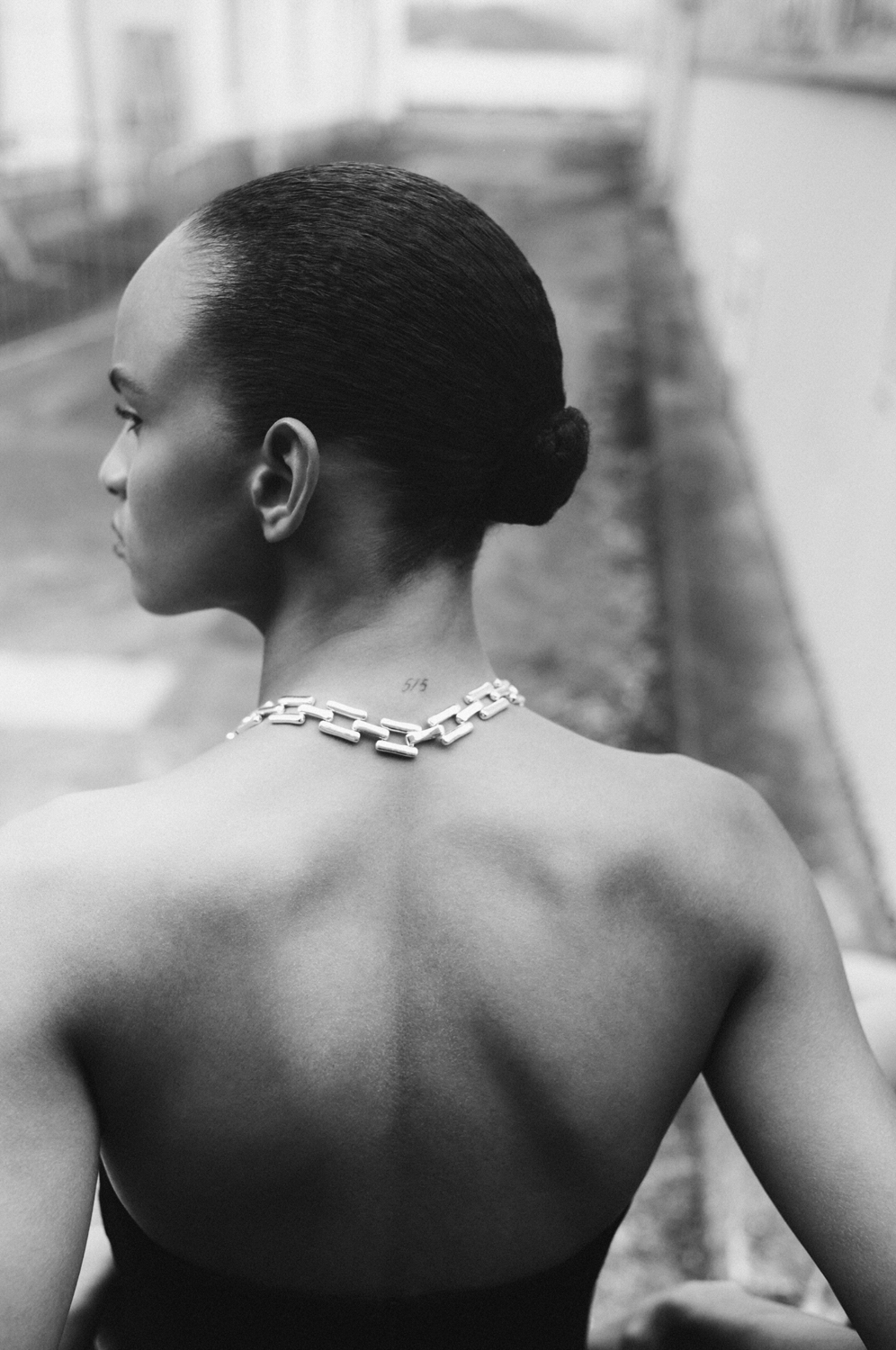 Monochrome portrait from behind of a woman with bare back, wearing a chain necklace, hair up, gazing to the left.