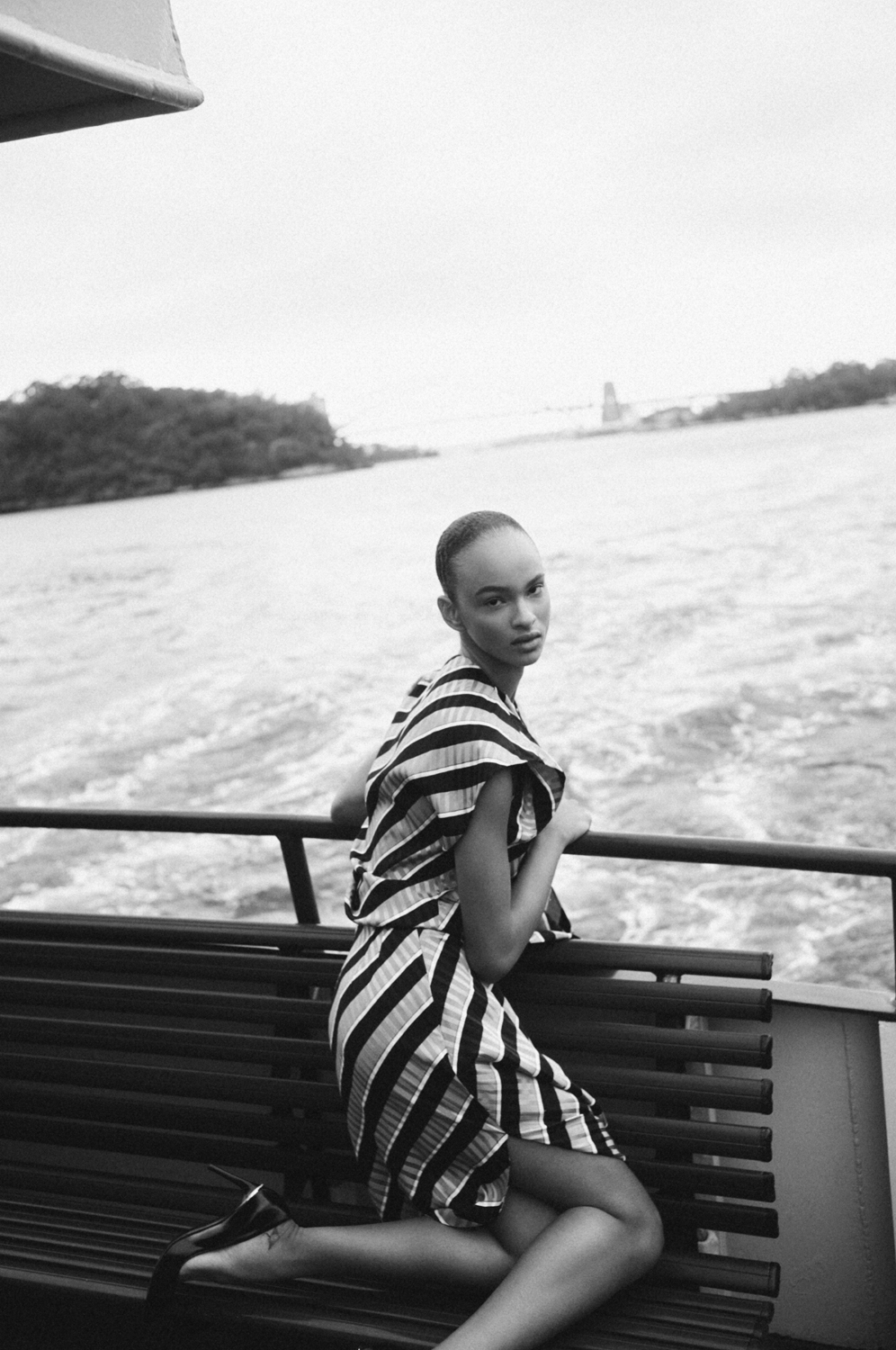 Black and white portrait of a model in a striped dress kneeling on a bench by the water with a distant city skyline.