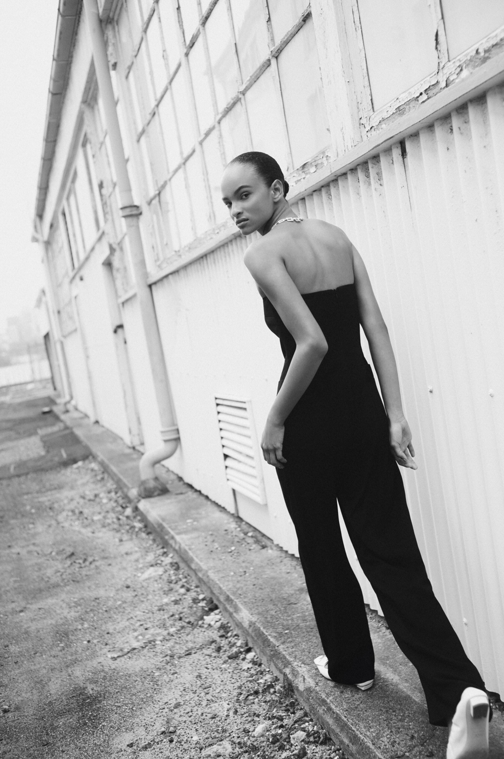A slender woman in a black jumpsuit walks on a concrete ledge in an urban setting, glancing over her shoulder in black and white.