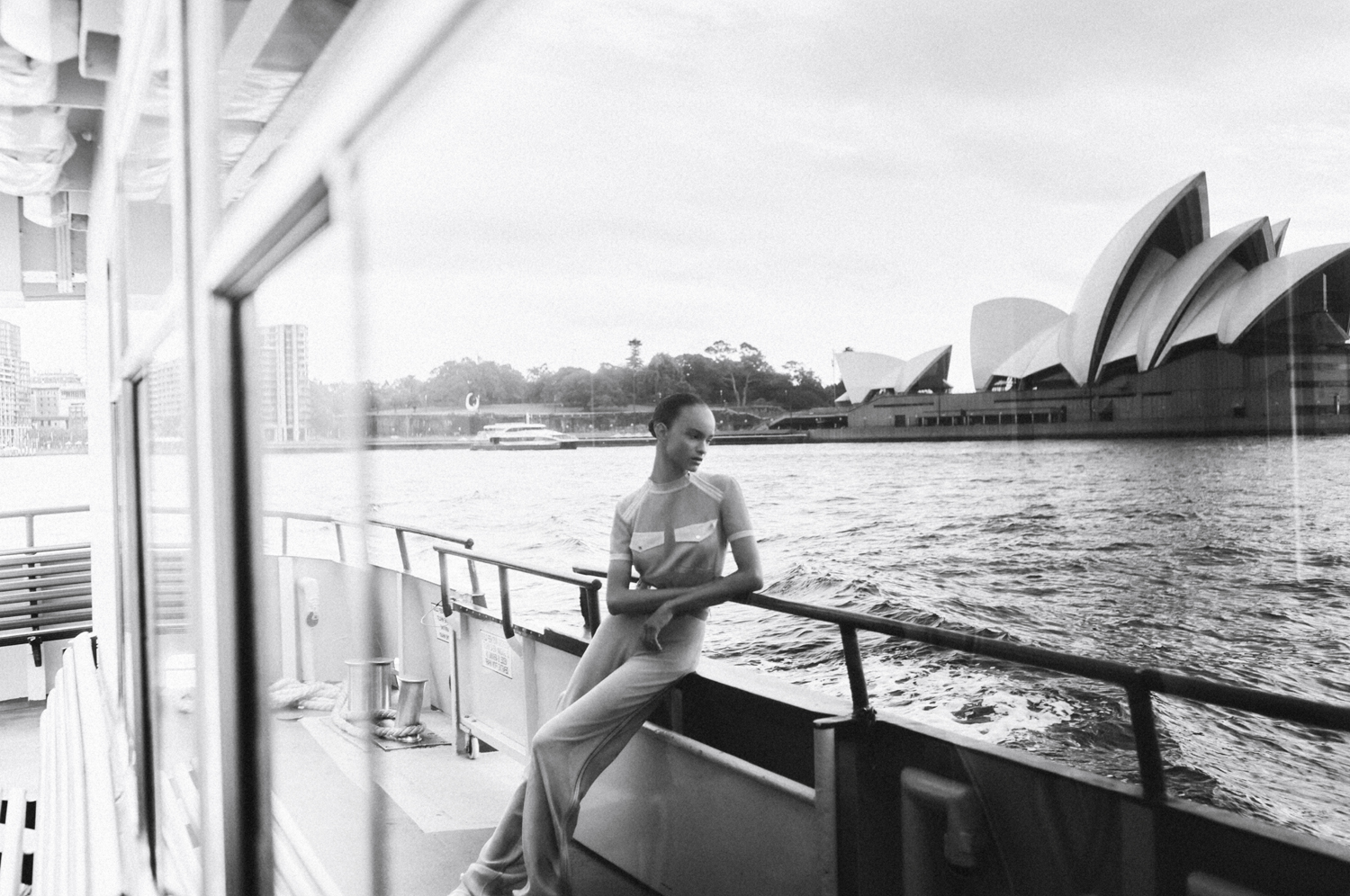 Monochrome shot of a fashion model in a jumpsuit reclining on the railing of a boat with the Sydney Opera House in the background.