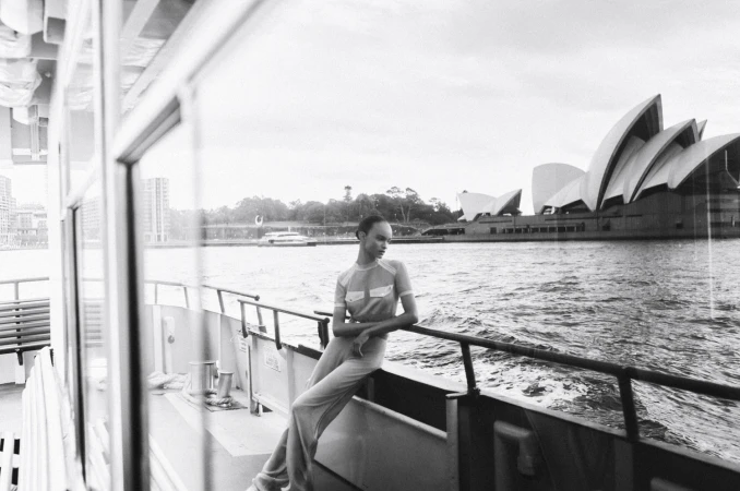 Monochrome shot of a fashion model in a jumpsuit reclining on the railing of a boat with the Sydney Opera House in the background.