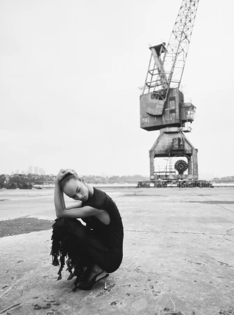 Woman in black dress poses near harbor crane. Moody black-and-white fashion shot with urban industrial backdrop and architectural elements.