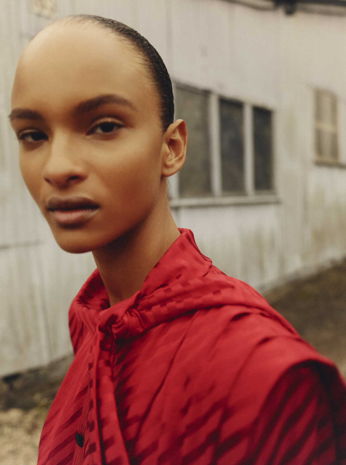 Close-up studio portrait of an adult woman with short dark hair, wearing a red patterned hooded jacket, against a blurred building backdrop.