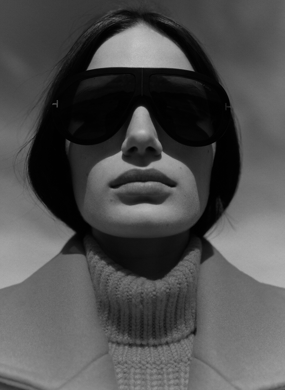 Close-up shot of a brunette woman wearing a beige coat, a turtleneck sweater, and oversized sunglasses with the brand logo visible.
