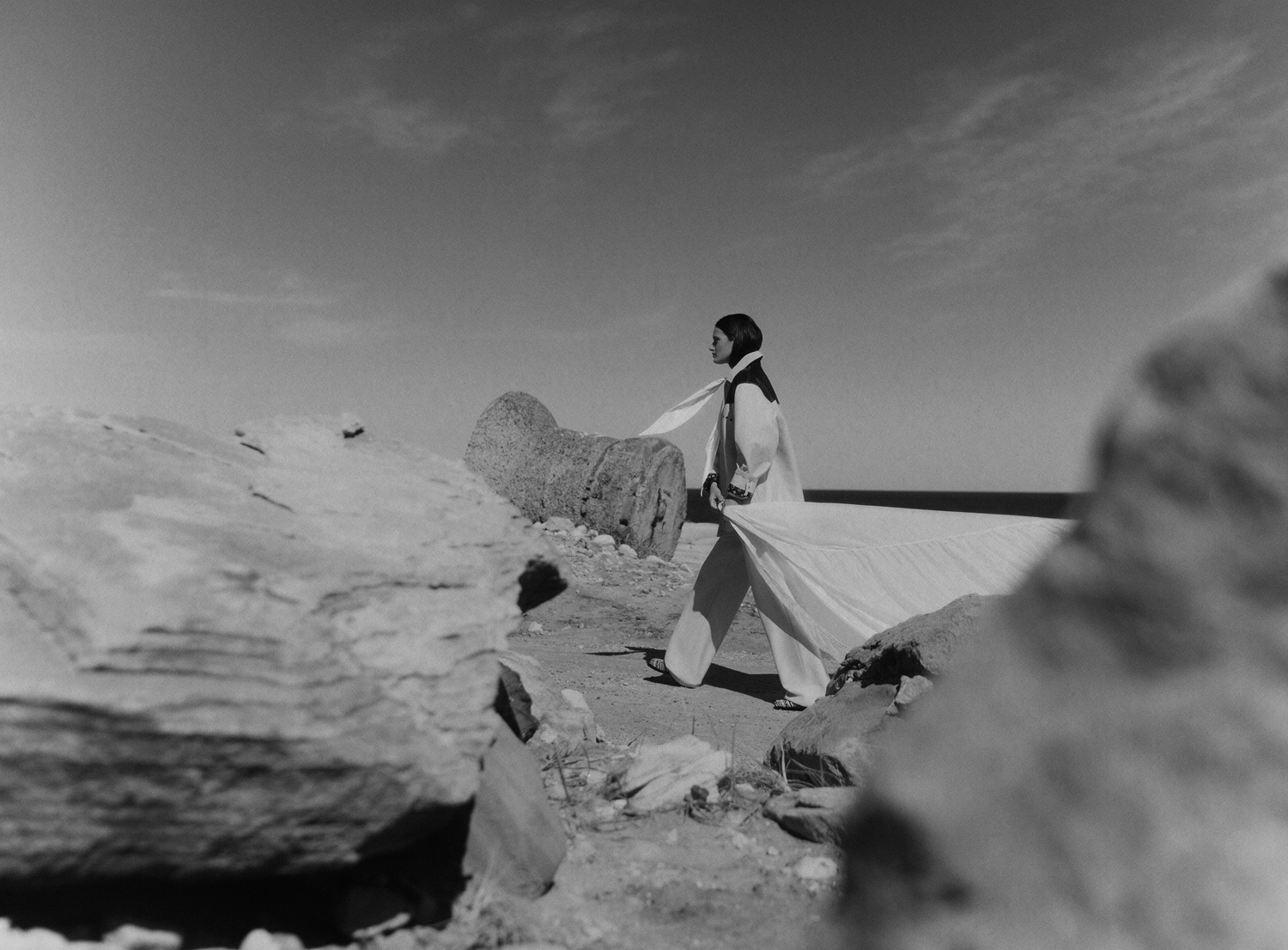 A woman walks on a rocky beach in a long white robe and scarf, with a black top, alongside remnants of stone columns in black and white.