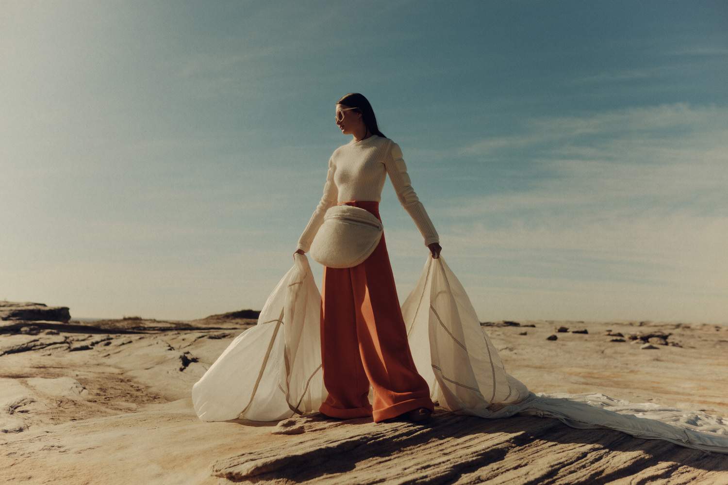 Woman in orange pants and white top holds a white fabric on a tan, rocky landscape under a bright sky, creating a minimalist fashion editorial.