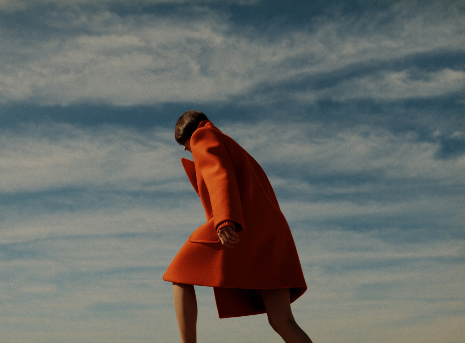 An adult female wearing a red coat poses outdoors, shot from a low angle against a cloudy blue sky, showcasing fashion and style.