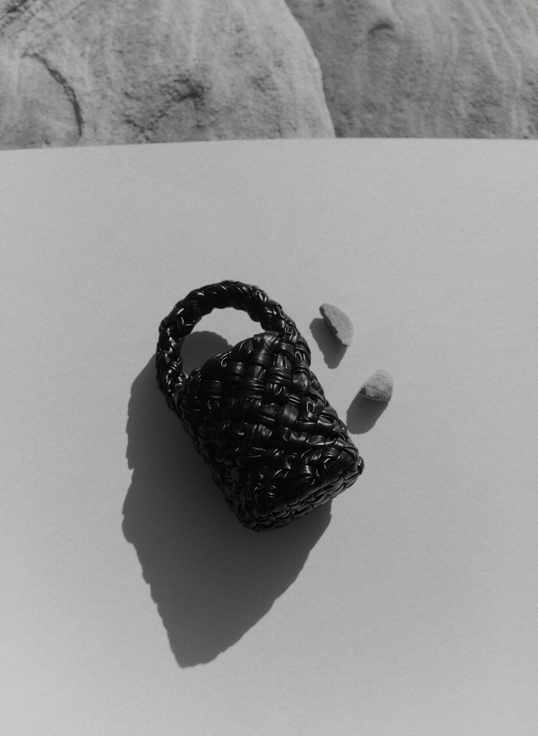 A black woven handbag with handle rests on a white surface with two small rocks in a studio shot with a monochrome color palette.