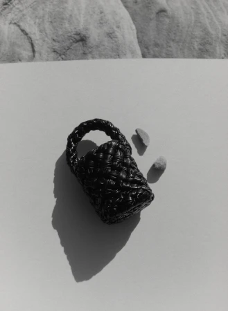 A black woven handbag with handle rests on a white surface with two small rocks in a studio shot with a monochrome color palette.