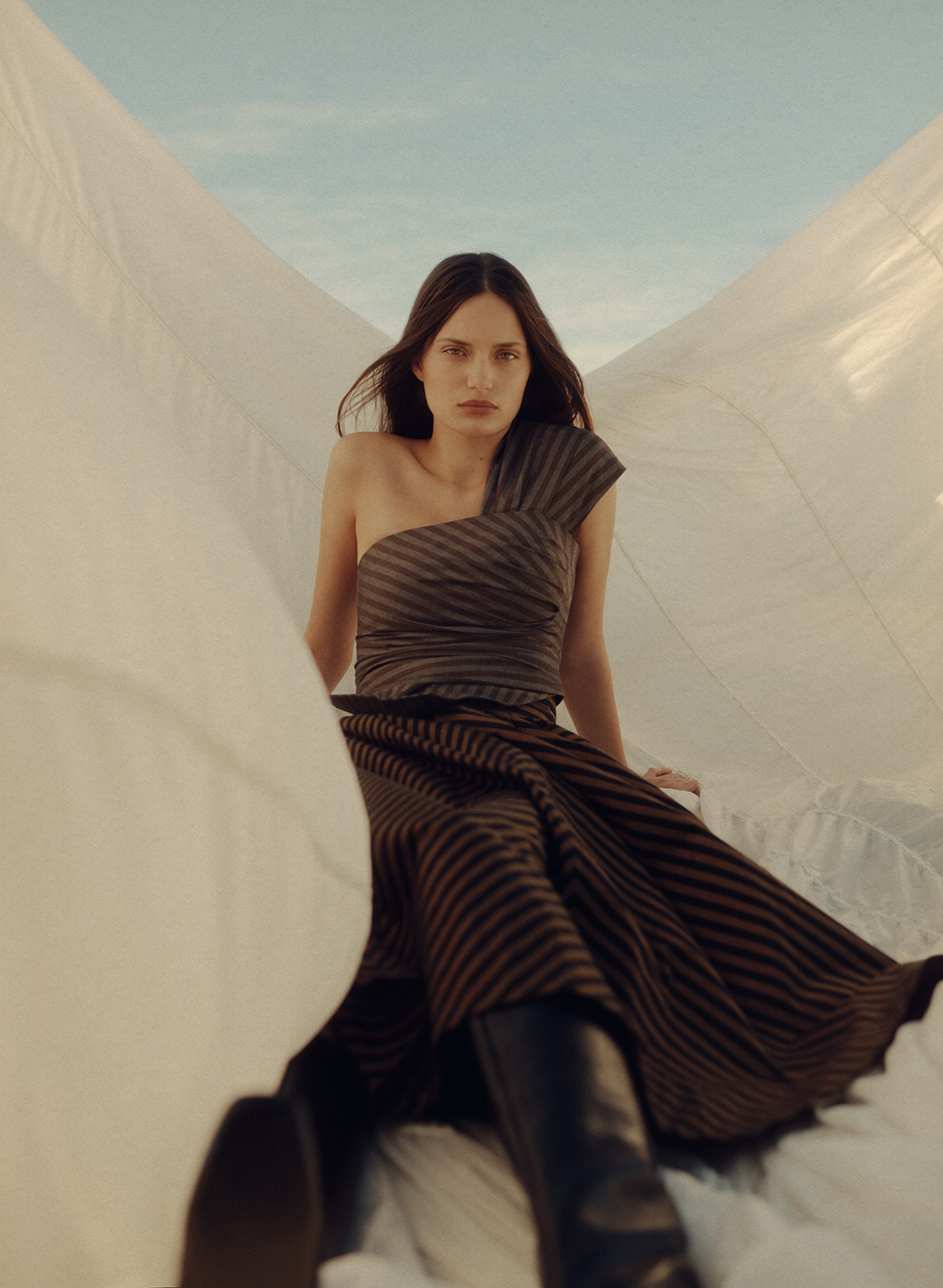 A woman in a brown striped one-shoulder dress and black boots poses against a white fabric backdrop under a blue sky for fashion.