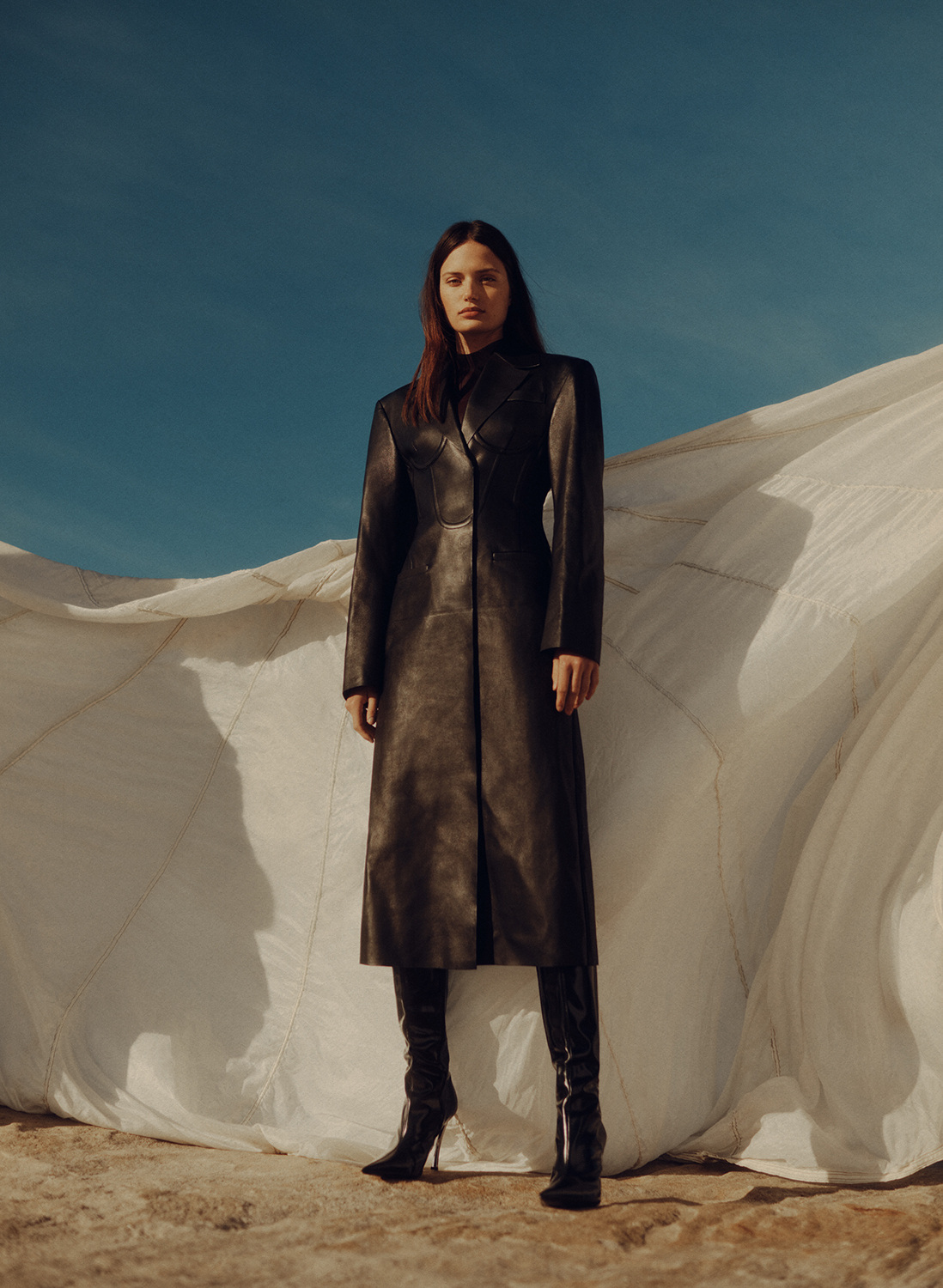 Model in a tailored long black leather coat and high-heeled boots posing in front of a draped white textile backdrop under clear sky.
