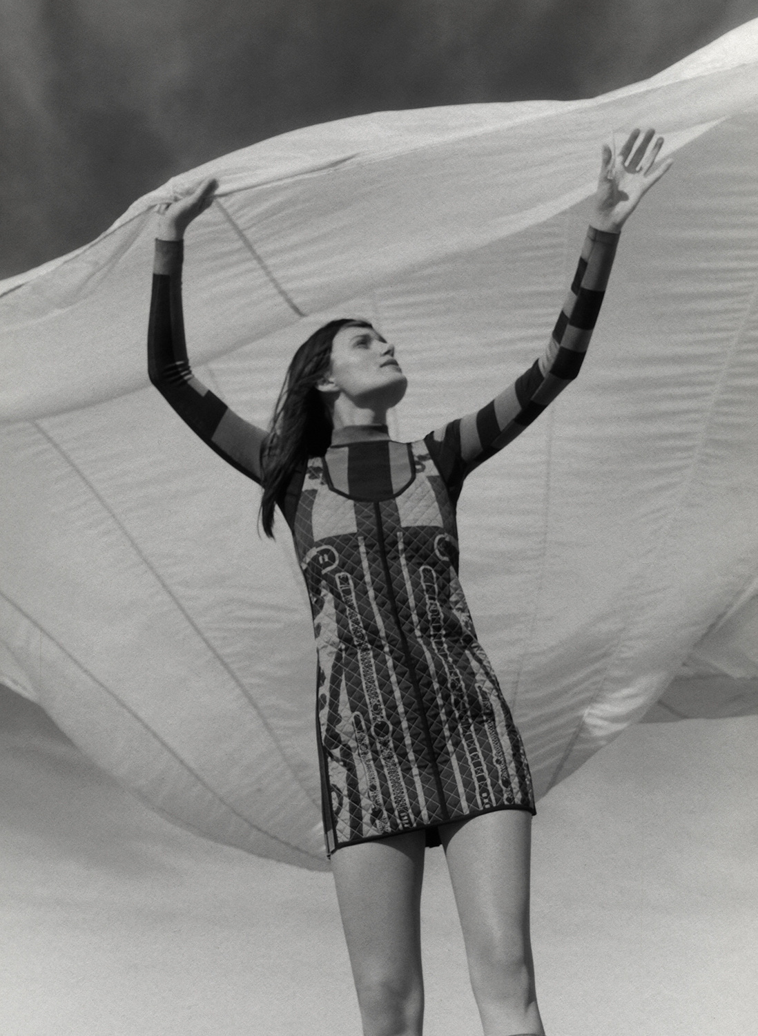 Monochrome fashion shot of a woman in patterned minidress under a light fabric, looking up with raised arms against a light sky.