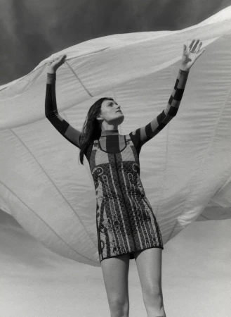 Monochrome fashion shot of a woman in patterned minidress under a light fabric, looking up with raised arms against a light sky.