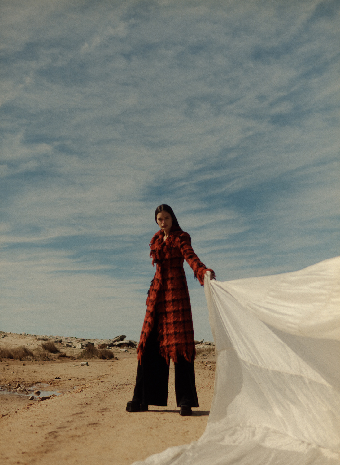 A woman in a red plaid coat stands on a beach holding a white sheet. The photo features cloudy skies, a tan sandy beach, and natural light.