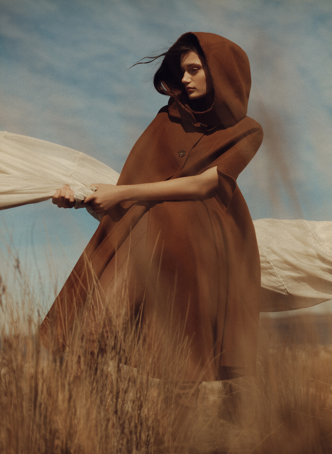 A woman in a brown hooded cape stands in a field of tall grass with a blue sky and clouds. Fashion style, outdoor setting.
