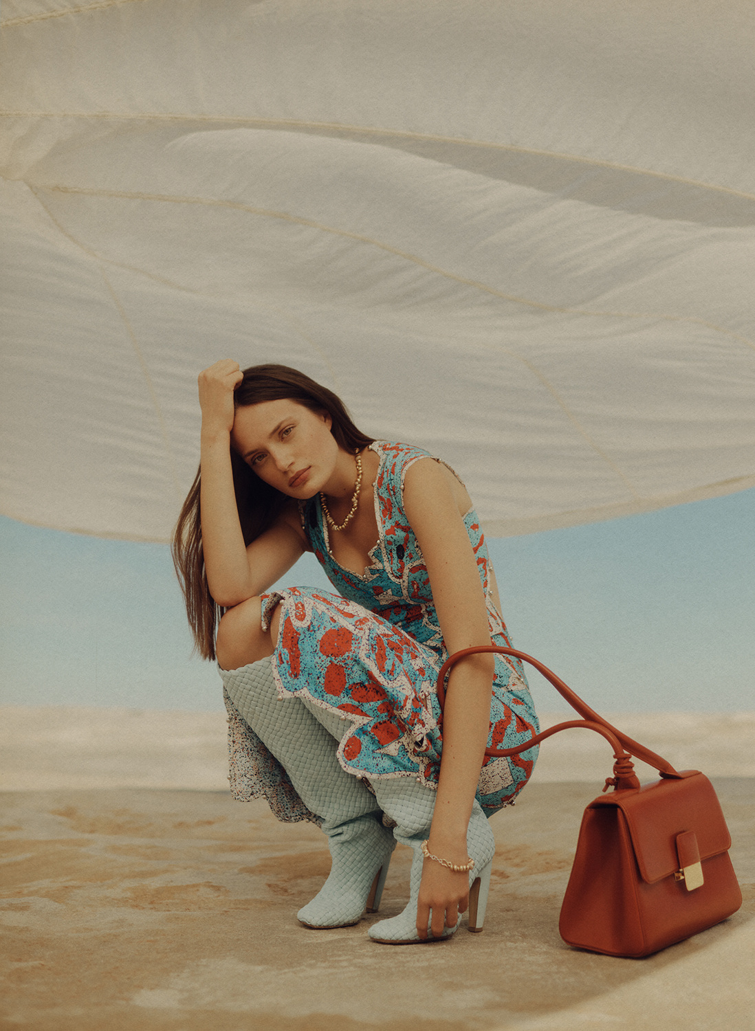 Woman in a floral dress and light blue boots crouching with a brown satchel bag in an outdoor setting with a blue sky backdrop.
