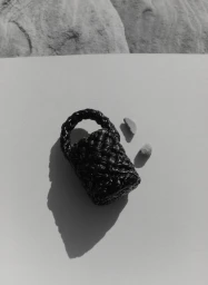 Monochrome still life of a miniature black woven leather handbag and two small stones casting sharp shadows on a pale surface.