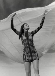 Model in patterned minidress holding up large white fabric sheet against sky