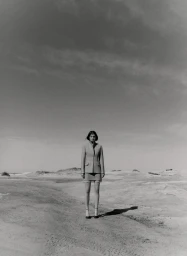 Model standing alone on a vast sandy landscape under a large overcast sky in a monochromatic editorial fashion photograph.