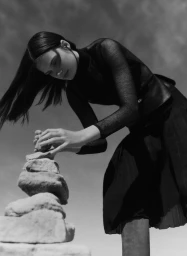 Model in a brown hooded cape holding white fabric in a grassy field under an open sky