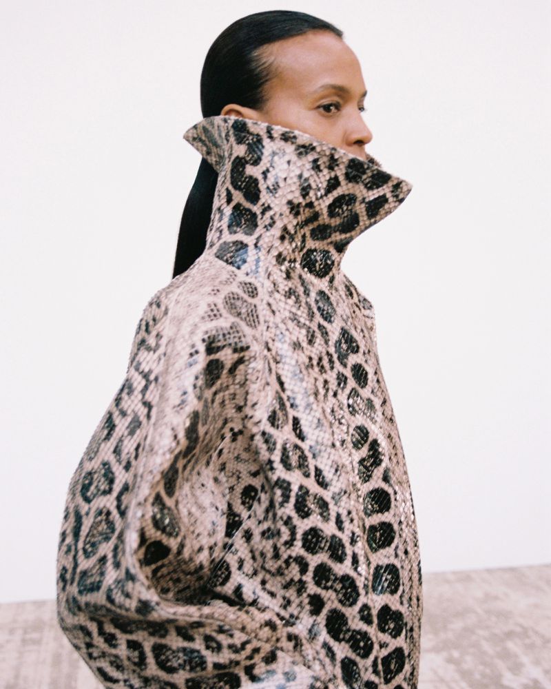 Model Liya Kebede in profile wearing a high-collared animal-print textured coat against a minimalist white studio backdrop.