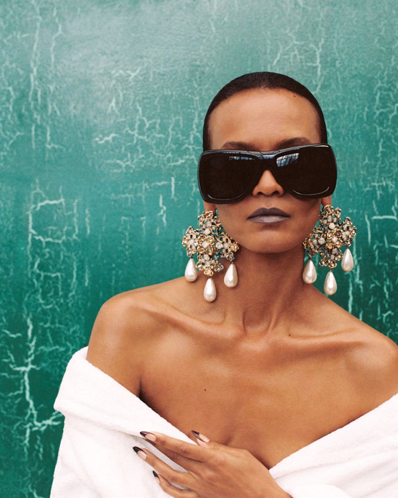 Model Liya Kebede wearing oversized black sunglasses and ornate pearl drop earrings against a green cracked backdrop.