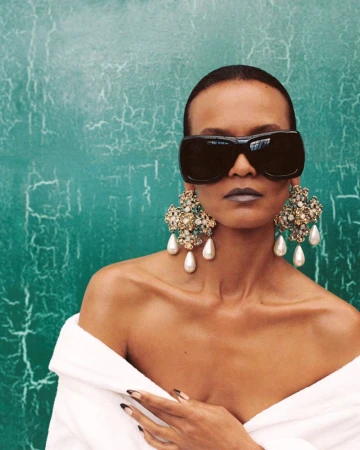 Model Liya Kebede wearing oversized black sunglasses and ornate pearl drop earrings against a green cracked backdrop.