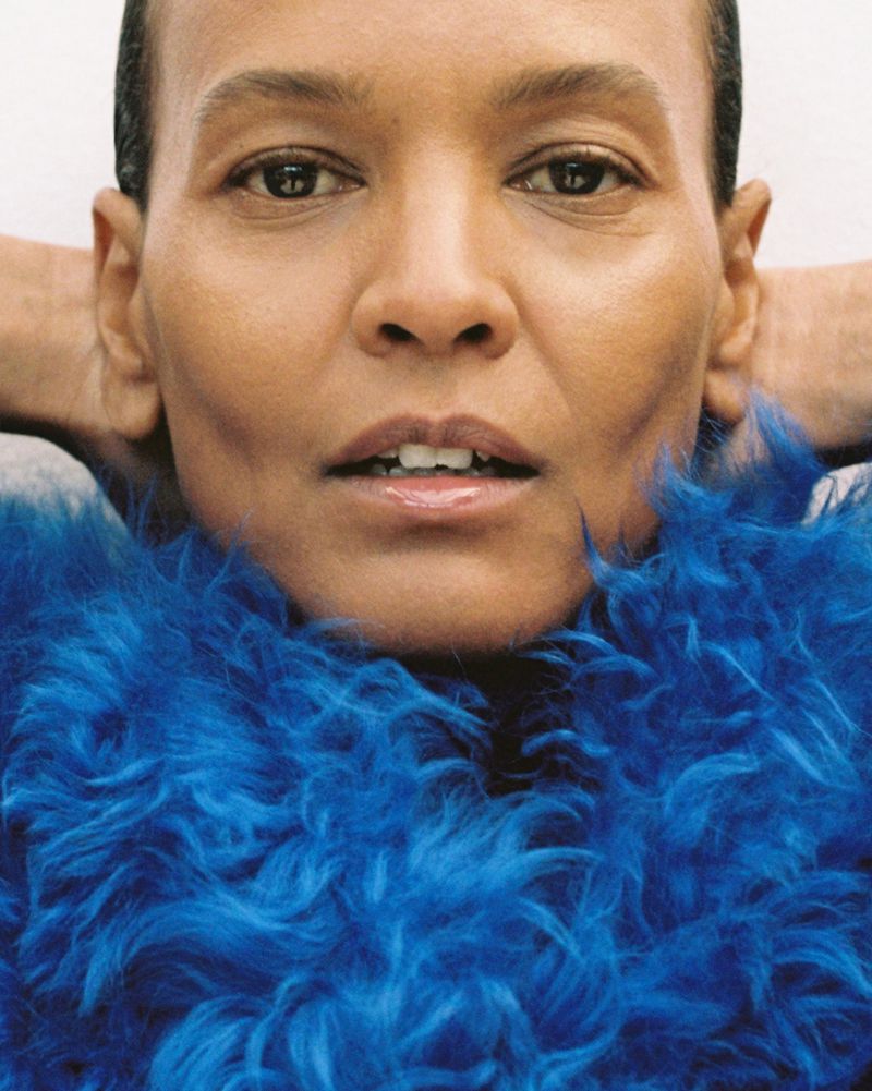 Extreme close-up portrait of model Liya Kebede wearing a bright cobalt blue textured faux fur collar with hands behind her head.