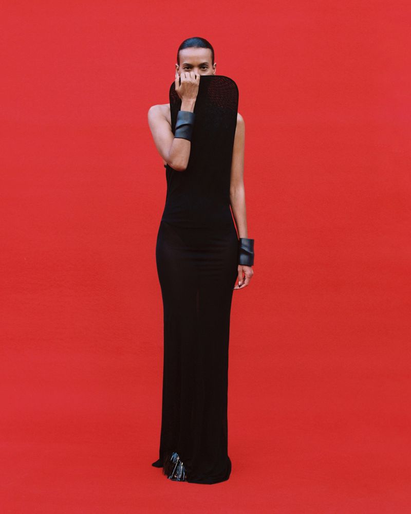 Model Liya Kebede standing in a sculptural black floor-length gown against a vivid red background.