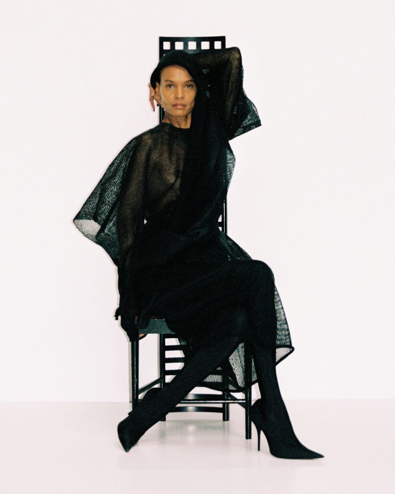 Model Liya Kebede seated on a dark high-backed chair in a textured dark mesh dress and heels against a white backdrop.