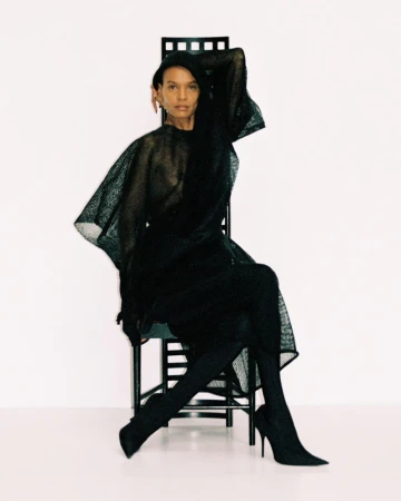 Model Liya Kebede seated on a dark high-backed chair in a textured dark mesh dress and heels against a white backdrop.