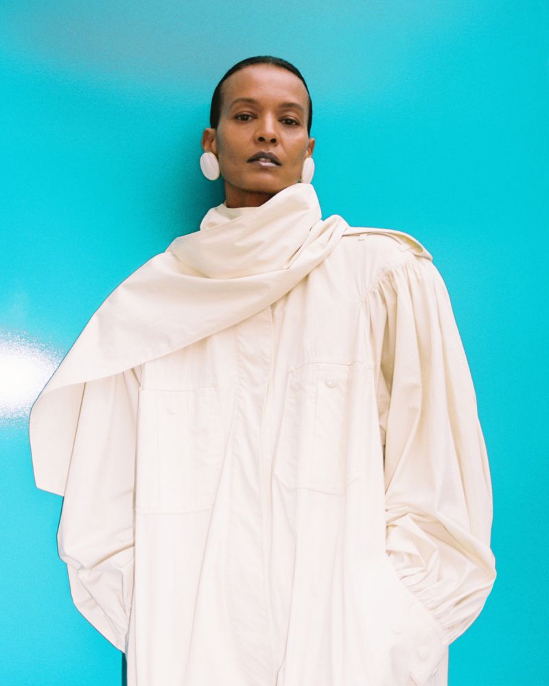 Liya Kebede wearing an oversized off-white draped coat and white disc earrings against a bright cyan blue backdrop.