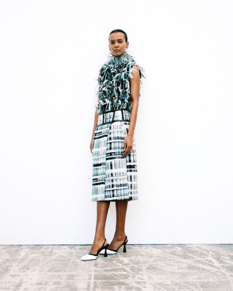 Model Liya Kebede standing in a textured sleeveless feather top and printed midi skirt with slingback heels against a white backdrop.