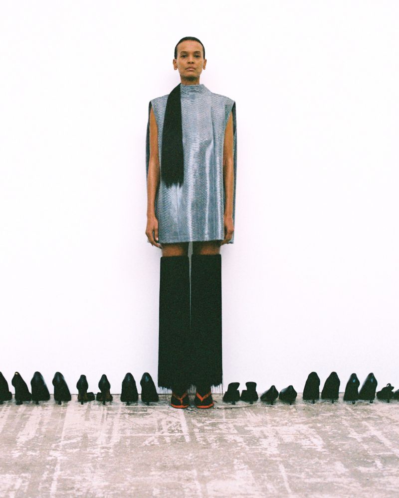 Liya Kebede standing in a silver sleeveless tunic and long black fringe against a white backdrop lined with black high heels.
