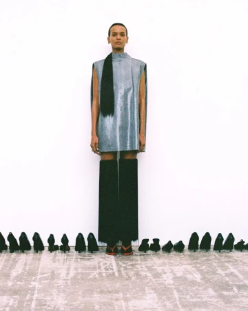 Liya Kebede standing in a silver sleeveless tunic and long black fringe against a white backdrop lined with black high heels.