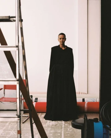 Model Liya Kebede posing in a voluminous black gown in a photo studio with a ladder and background rolls visible.