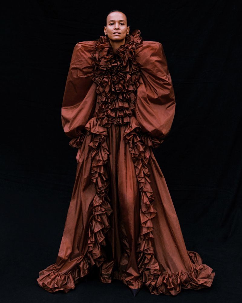Liya Kebede standing in an exaggerated ruffled brown gown with voluminous puffed shoulders against a black background.