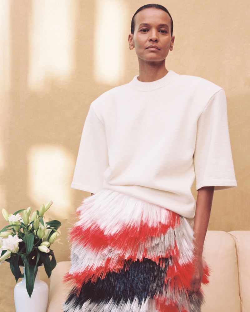 Liya Kebede posing in a cream structured top and a tiered red, black, and white fringe skirt beside a white vase of flowers.