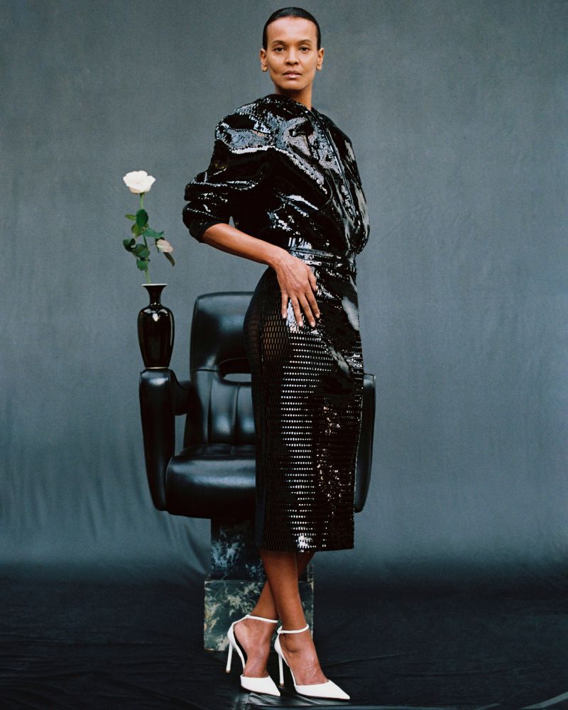 Liya Kebede standing beside a black leather armchair and a single white rose in a black vase against a slate backdrop.