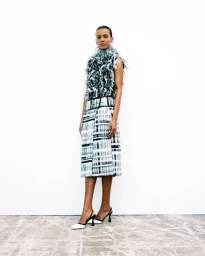 Model Liya Kebede standing in a textured sleeveless feather top and printed midi skirt with slingback heels against a white backdrop.